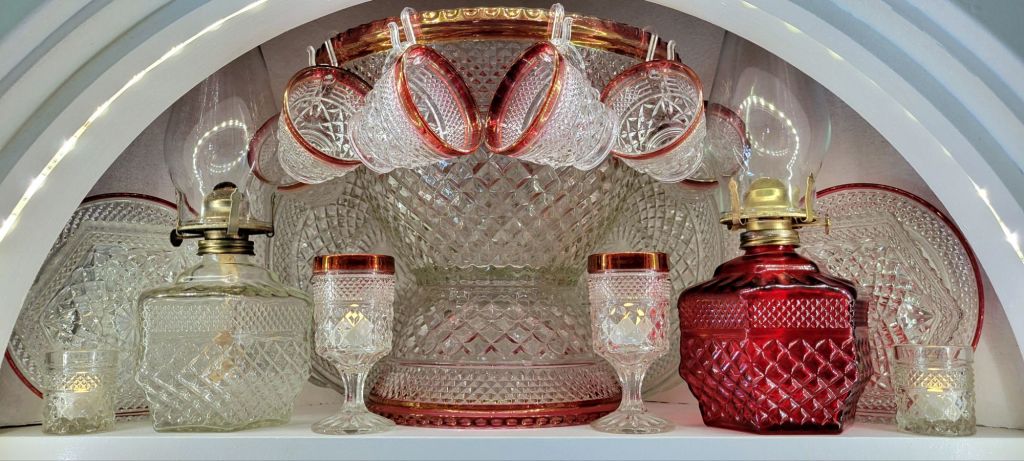 the top shelf of an open-doored, built-in hutch showing of it's contents of clear Wexford and ruby-banded Oxford glassware by Anchor Hocking, featuring a ruby-banded punch set, and large oil lamps.