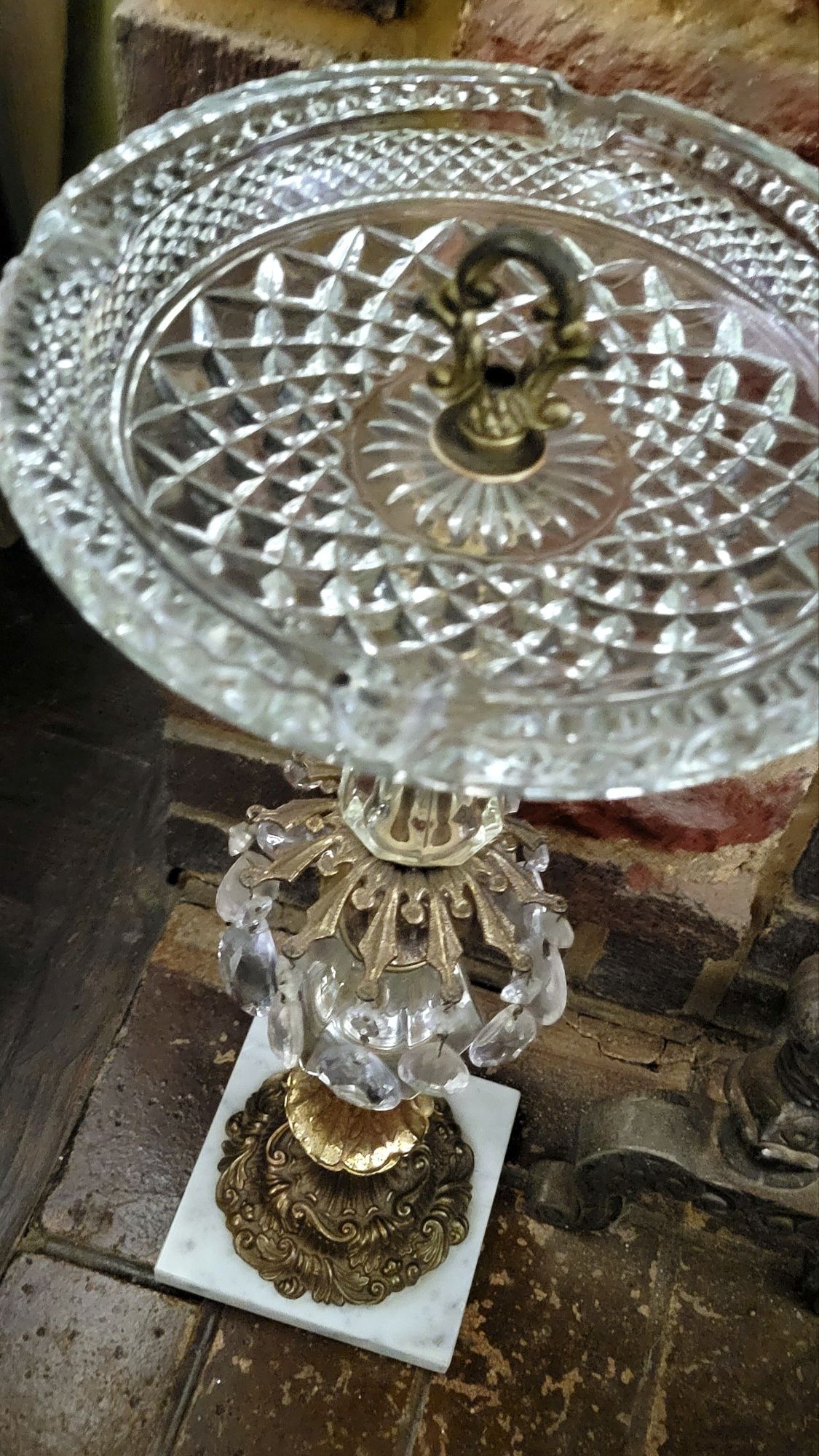 an angled overhead view of a Hollywood Regency-style ashtray stand, showing details of the marble base, brass segments, and crystal chandelier baubles. 