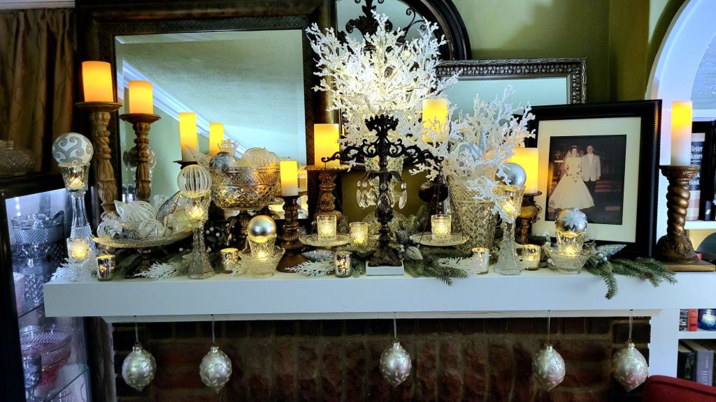 a mantle dressed for the holidays: chunky gilded candlesticks support LED pillar candles, Wexford clear pressed glass pieces fill in the remaining spaces, some used in Hollywood Regency-style assembled pieces. artificial winter branches are spread underneath and fill two tall vases; and clear, white, and silver glass ornaments fill the vessels. various votives dance with the glint of LED tealights. large ornate ornaments hang below the mantle. 