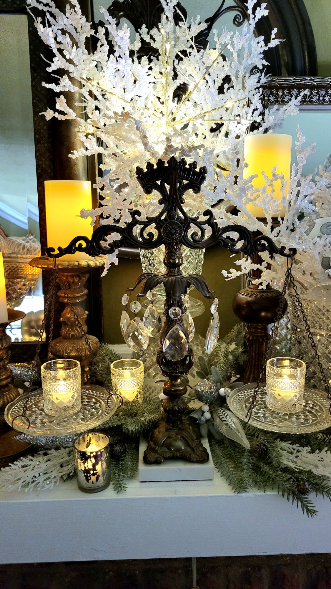 a mantle dressed for the holidays: chunky gilded candlesticks support LED pillar candles, Wexford clear pressed glass pieces fill in the remaining spaces, some used in Hollywood Regency-style assembled pieces. artificial winter branches are spread underneath and fill two tall vases; and clear, white, and silver glass ornaments fill the vessels. various votives dance with the glint of LED tealights.