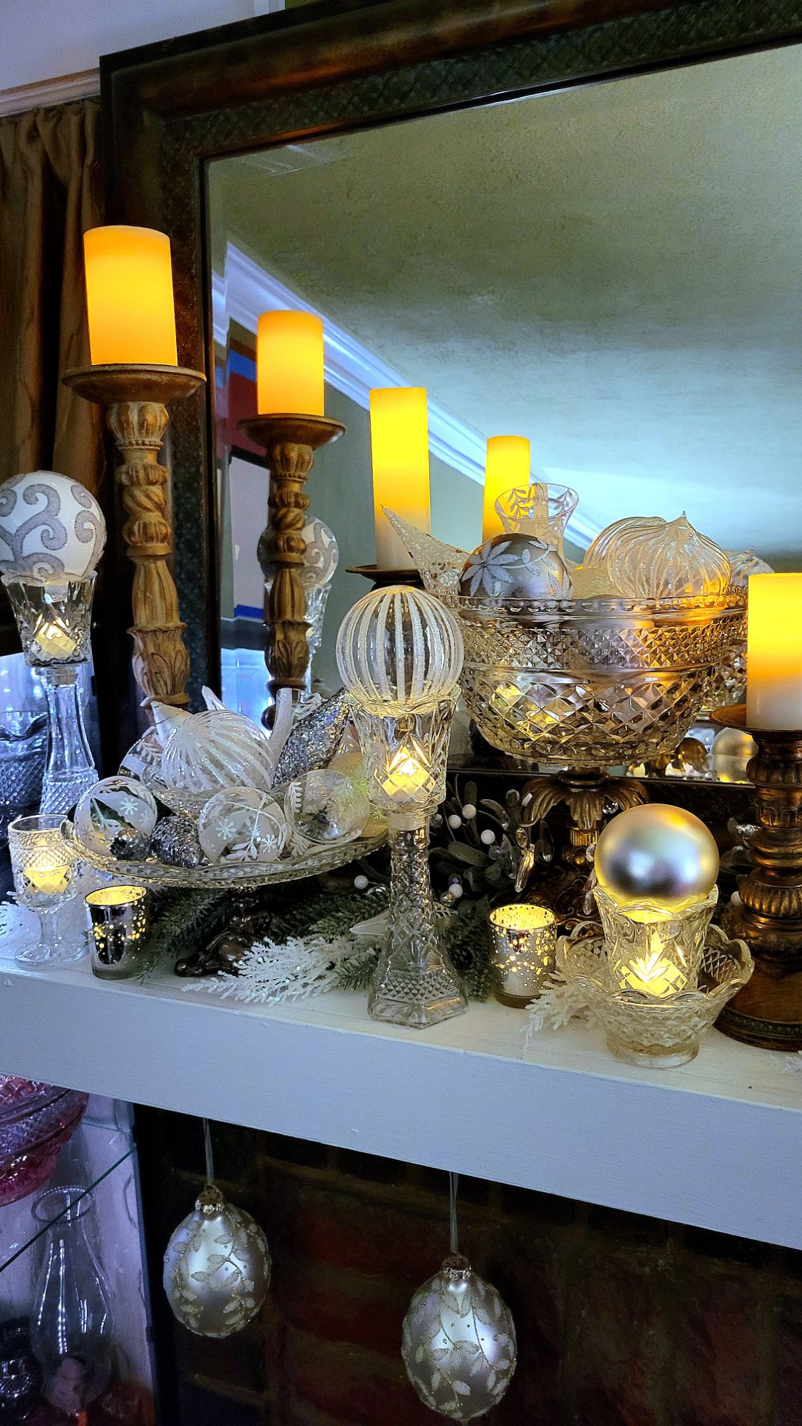 a mantle dressed for the holidays: chunky gilded candlesticks support LED pillar candles, Wexford clear pressed glass pieces fill in the remaining spaces, some used in Hollywood Regency-style assembled pieces. artificial winter branches are spread underneath and fill two tall vases; and clear, white, and silver glass ornaments fill the vessels. various votives dance with the glint of LED tealights. large ornate ornaments hang below the mantle. 