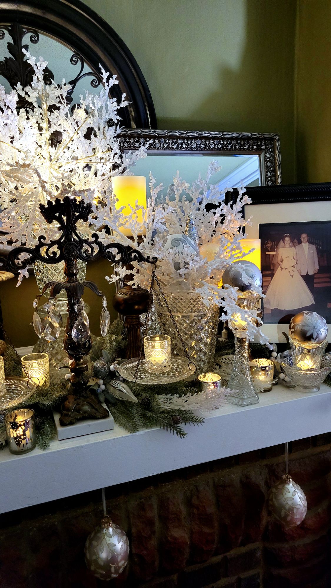 a mantle dressed for the holidays: chunky gilded candlesticks support LED pillar candles, Wexford clear pressed glass pieces fill in the remaining spaces, some used in Hollywood Regency-style assembled pieces. artificial winter branches are spread underneath and fill two tall vases; and clear, white, and silver glass ornaments fill the vessels. various votives dance with the glint of LED tealights. large ornate ornaments hang below the mantle. 