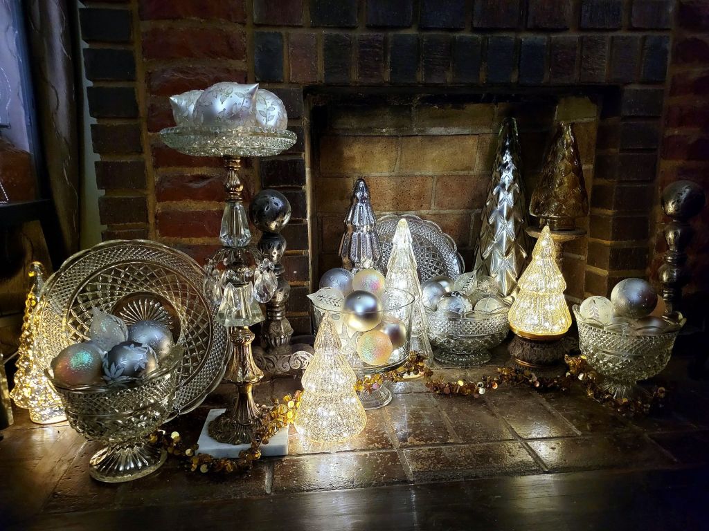 the fireplace box, overflowing with Wexford glass vessels filled with clear, white, and silver glass ornaments. just outside the opening is an elaborate Regency-style ashtray stand dripping with glass baubles and filled with large ornate glass ornaments. 