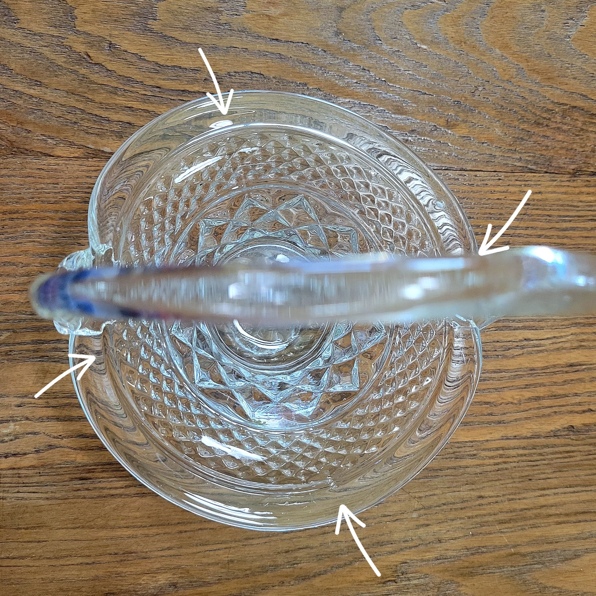 an overhead view of a pressed glass basket base, mimicking Wexford by Anchor Hocking, with a bent glass handle added presumably through a glassblowing technique. the arrows indicate the mold seams of the basket base and shows the off-center placement of the handle.