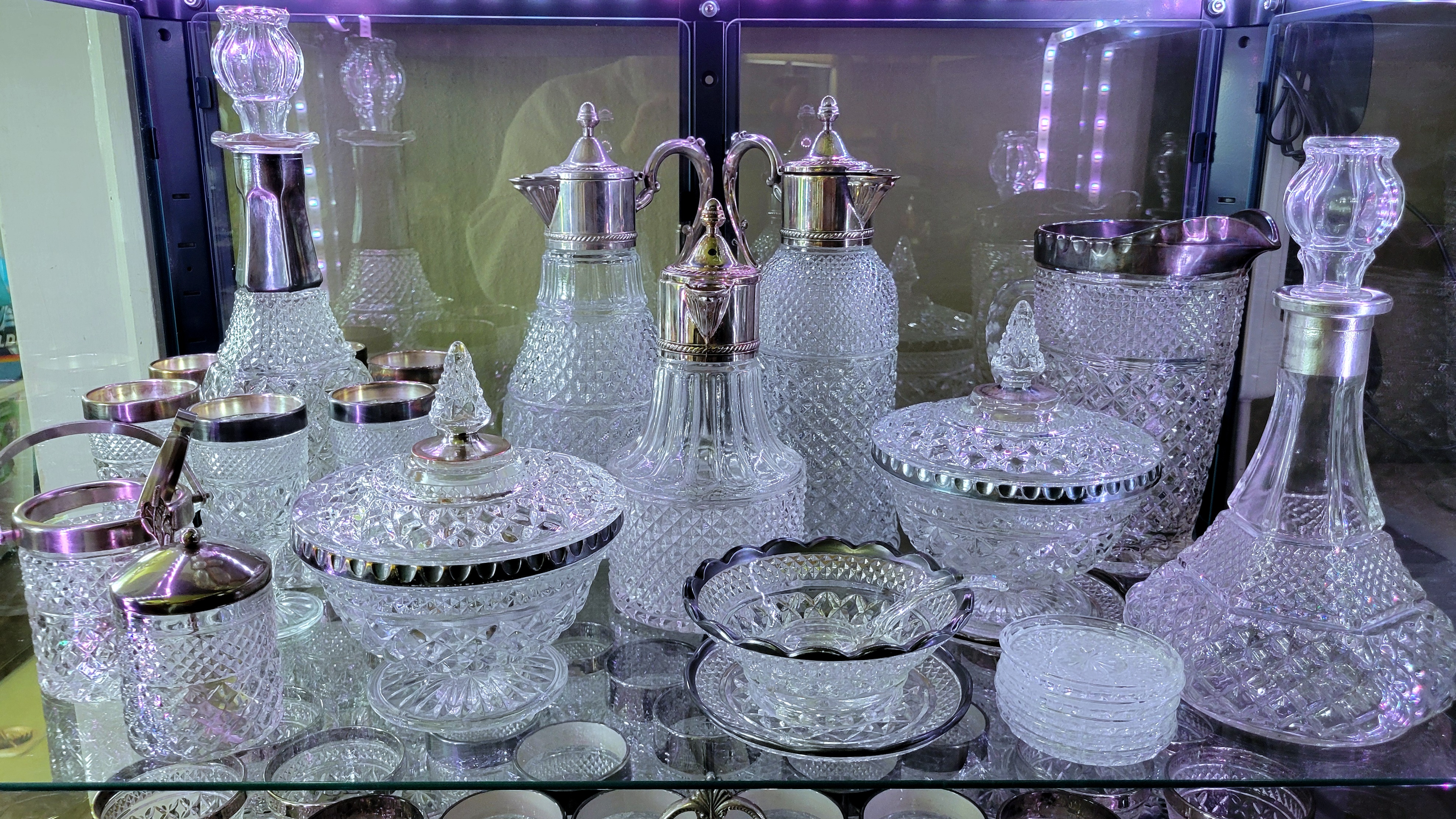 another display shelf of mostly tarnished silver-trimmed Wexford by Anchor Hocking. some European lookalike lidded pitchers, lookalike lead crystal coasters, and jam server, and sugar pail join the mix.