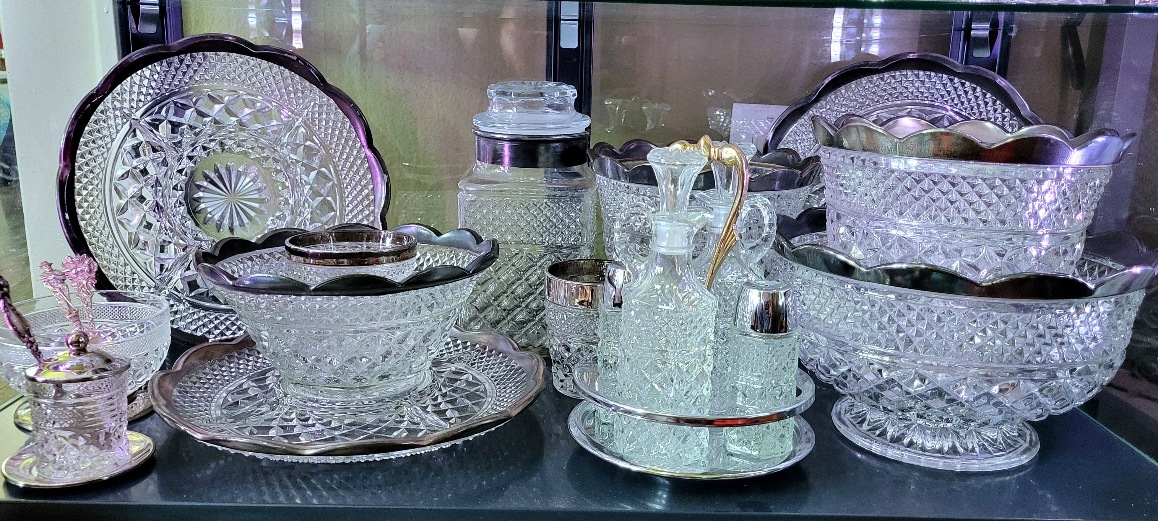 a display shelf of mostly heavily-tarnished silver trimmed Wexford by Anchor Hocking. a lookalike footed dish, a third-party jam serving using a Wexford votive holder, and a Kromex Vouge caddy holding regular crystal Wexford are mixed in.
