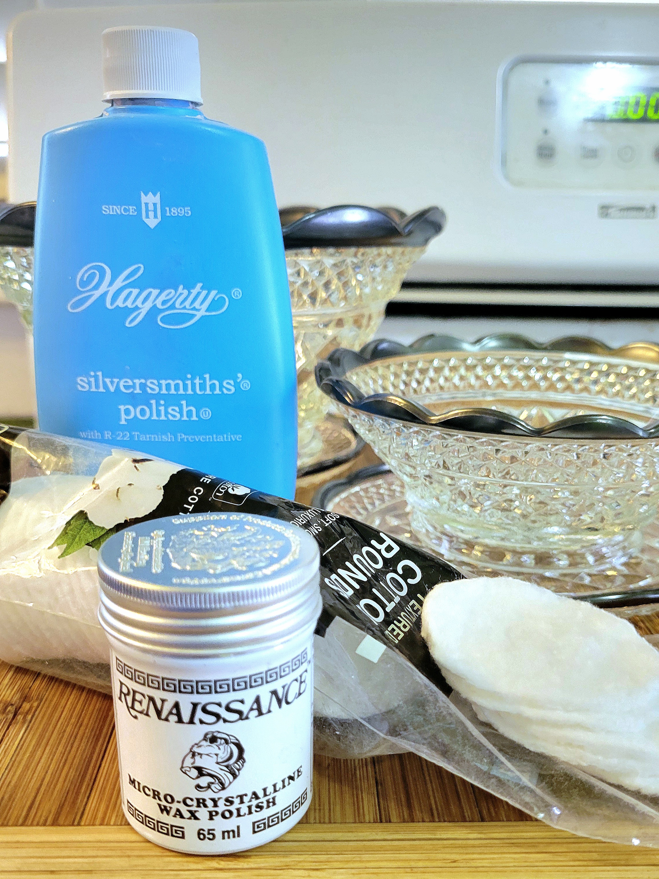 a still life of Hagerty silversmith polish, a tin of Renaissance micro-crystalline wax polish, cotton makeup pads, and some tarnished silver-trimmed scalloped edged Wexford pieces on the stovetop, waiting for the process to begin.