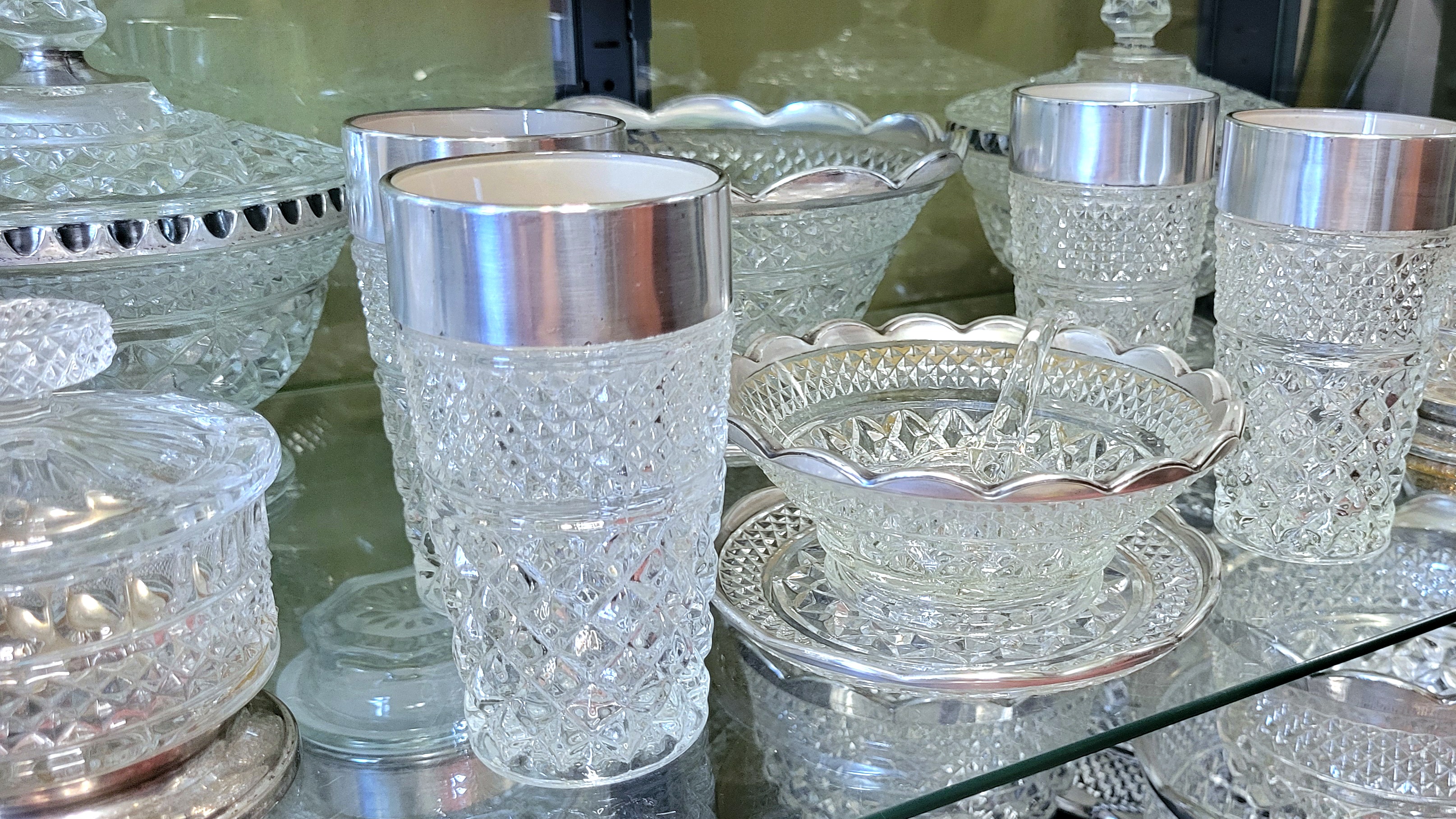 the middle display shelf, with smaller Wexford serving pieces and glassware. to the back left is a lidded candy dish with the patina left on the recesses of the silver trim to avoid rubbing the edges bare. to the back left is a tight view of the candy dish left unpolished due to silver loss.