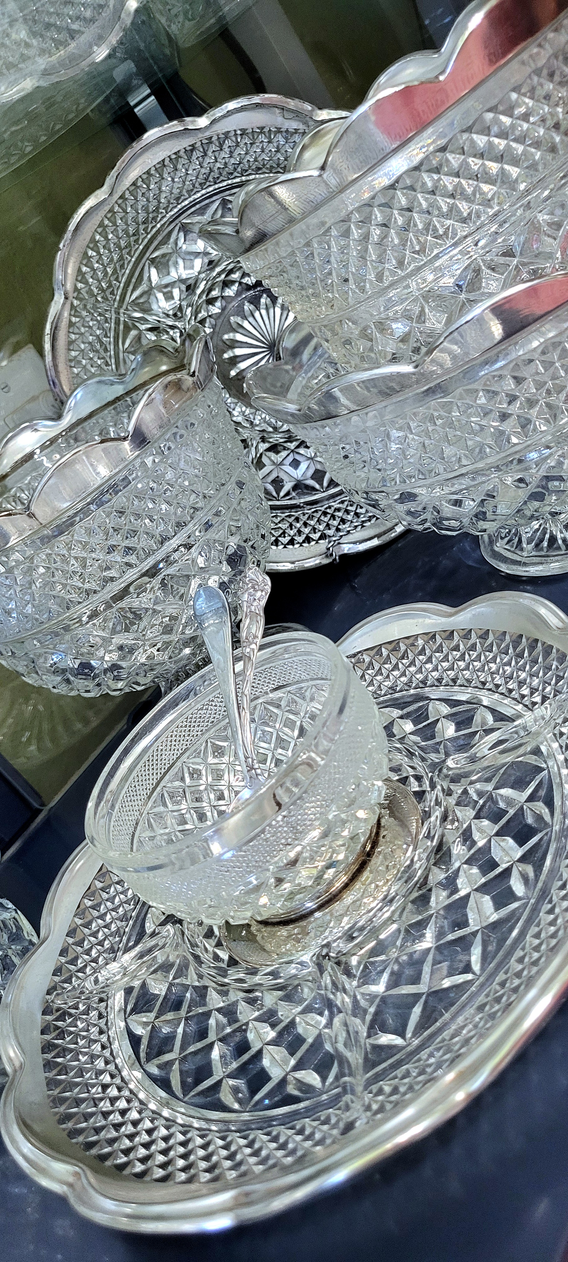 a close-up of the bottom shelf, witht he larger silver-trimmed serving pieces. the silver-trimmed scalopped edges light up the case with bouncing reflections.