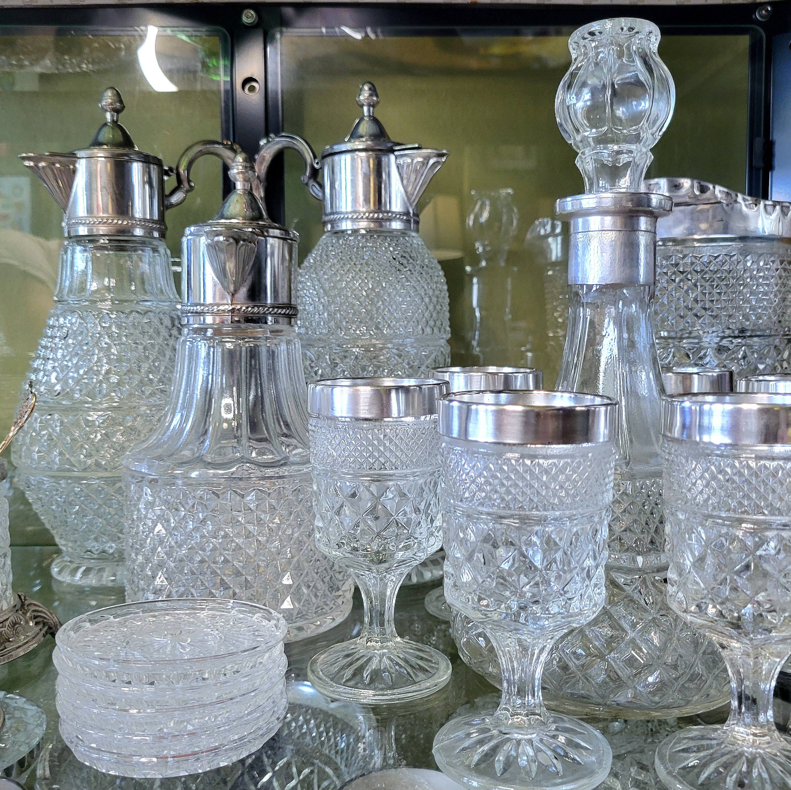 a close-up of the top shelf, full of pitchers, decanters, and glasses, all with silver trim. a stack of lookalike lead crystal coasters set in the front of all of it.
