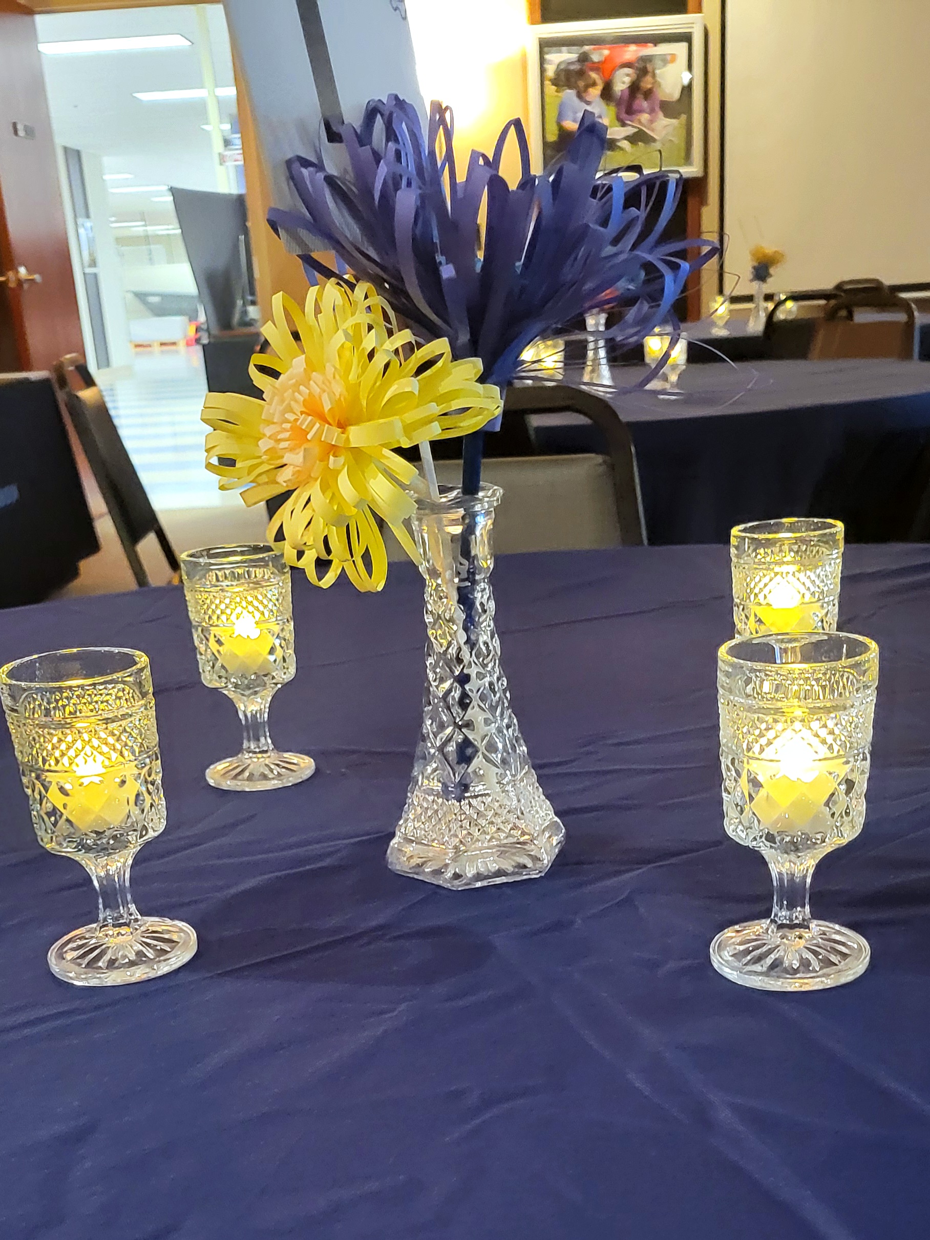 pressed glass cordial glasses lit with LED tealights surround a glass bud vase with paper flowers and a table number.