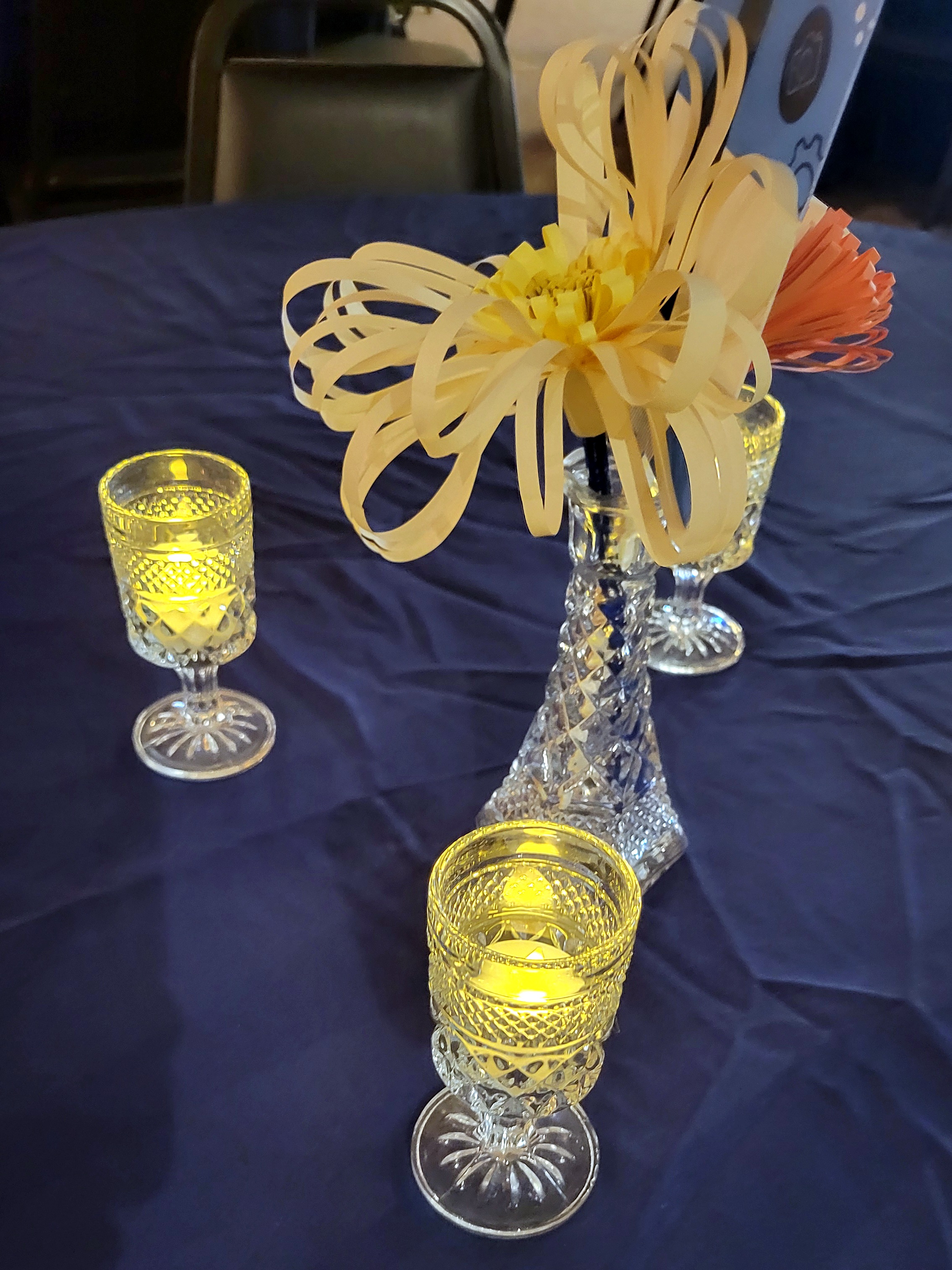 pressed glass cordial glasses lit with LED tealights surround a glass bud vase with paper flowers and a table number.