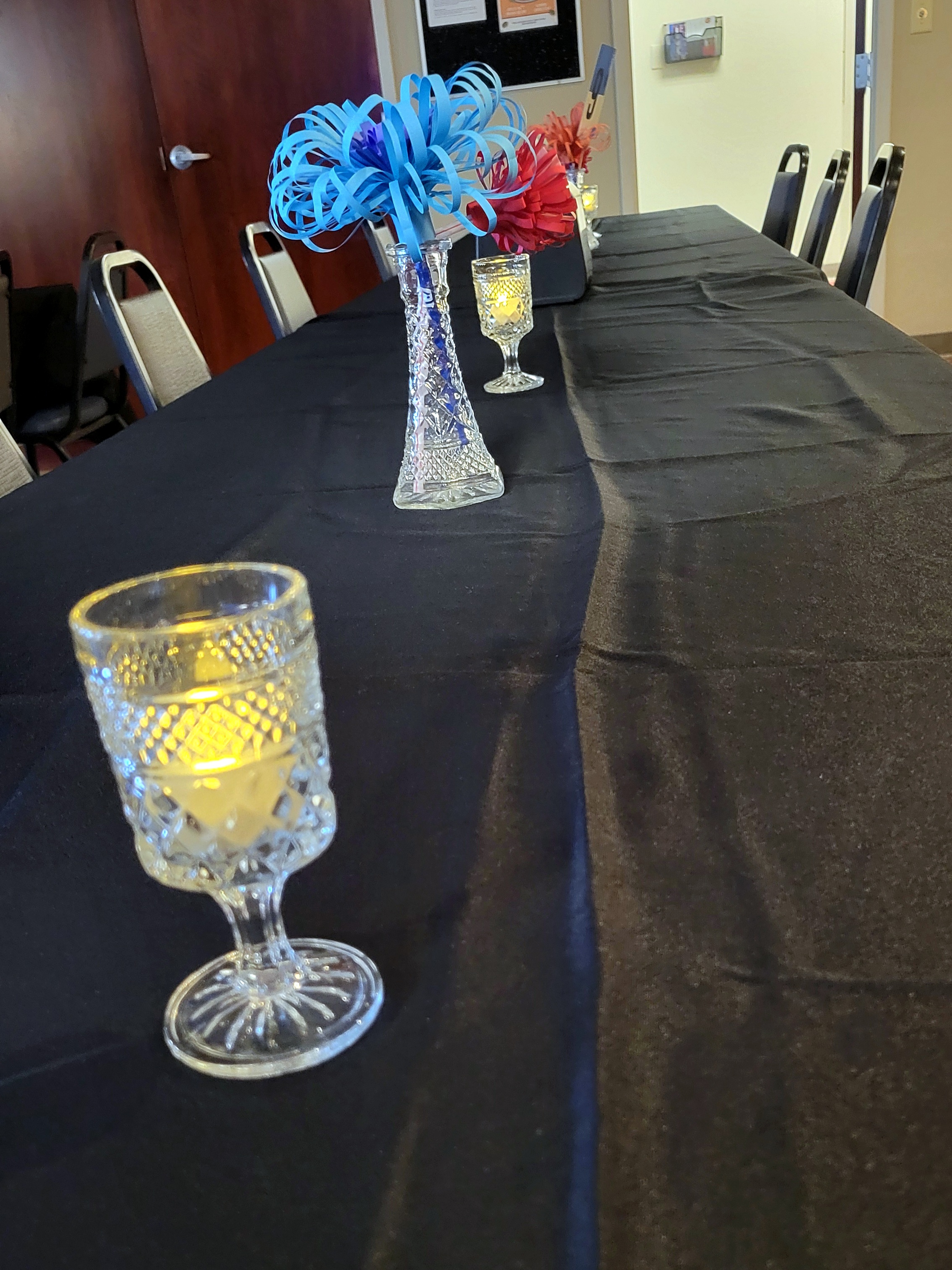 the same cordial glasses with tealights line up with glass bud vases holding paper flowers on a long rectangular table.