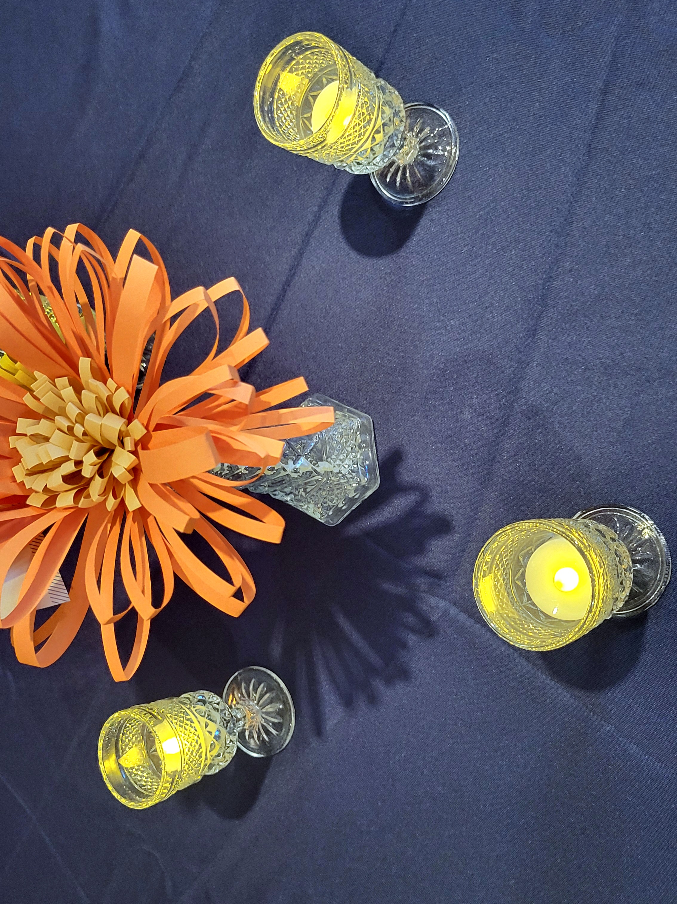 pressed glass cordial glasses lit with LED tealights surround a glass bud vase with paper flowers.