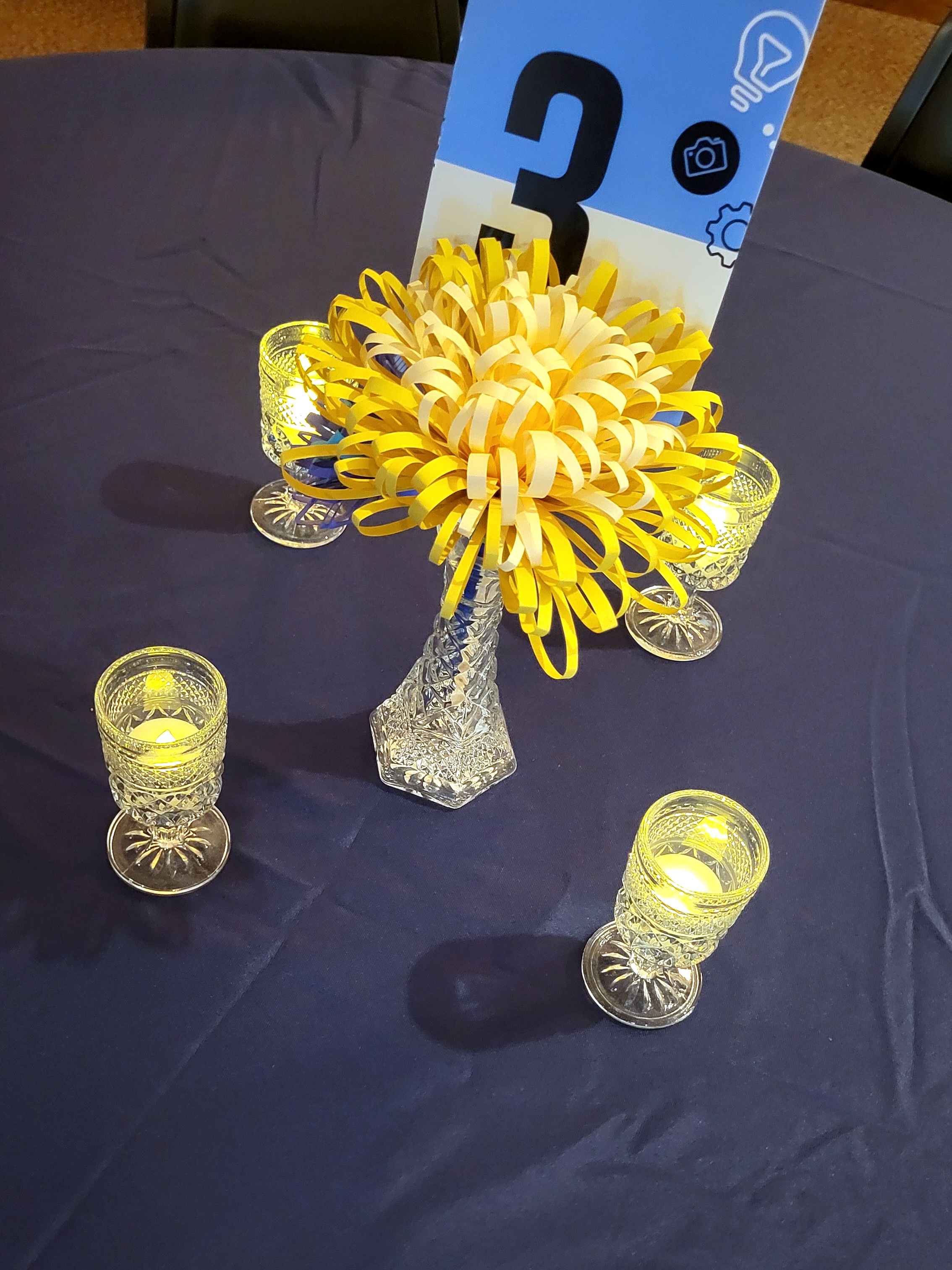 pressed glass cordial glasses lit with LED tealights surround a glass bud vase with paper flowers and a table number.