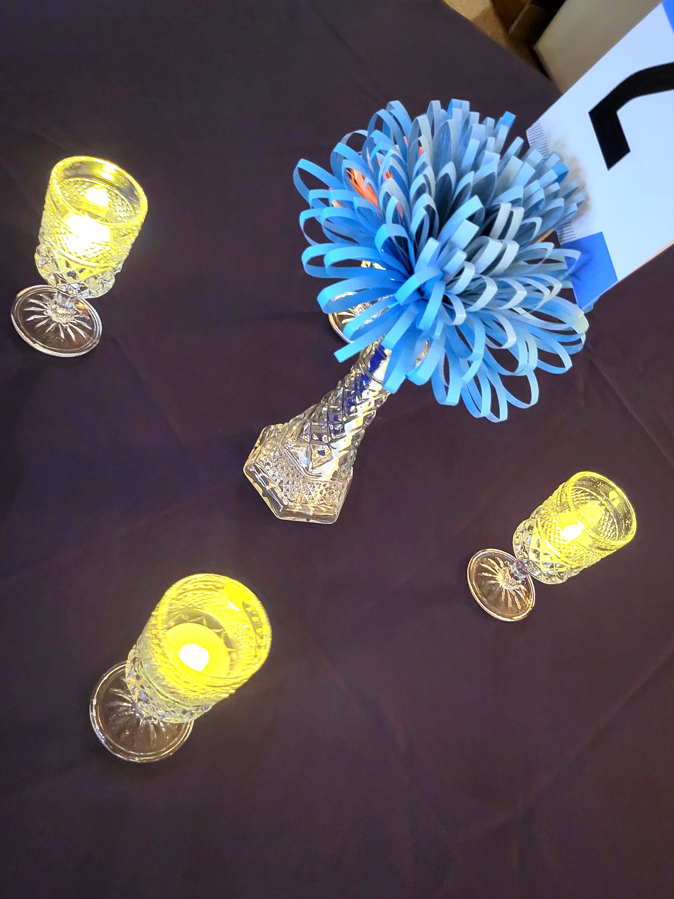 pressed glass cordial glasses lit with LED tealights surround a glass bud vase with paper flowers and a table number.