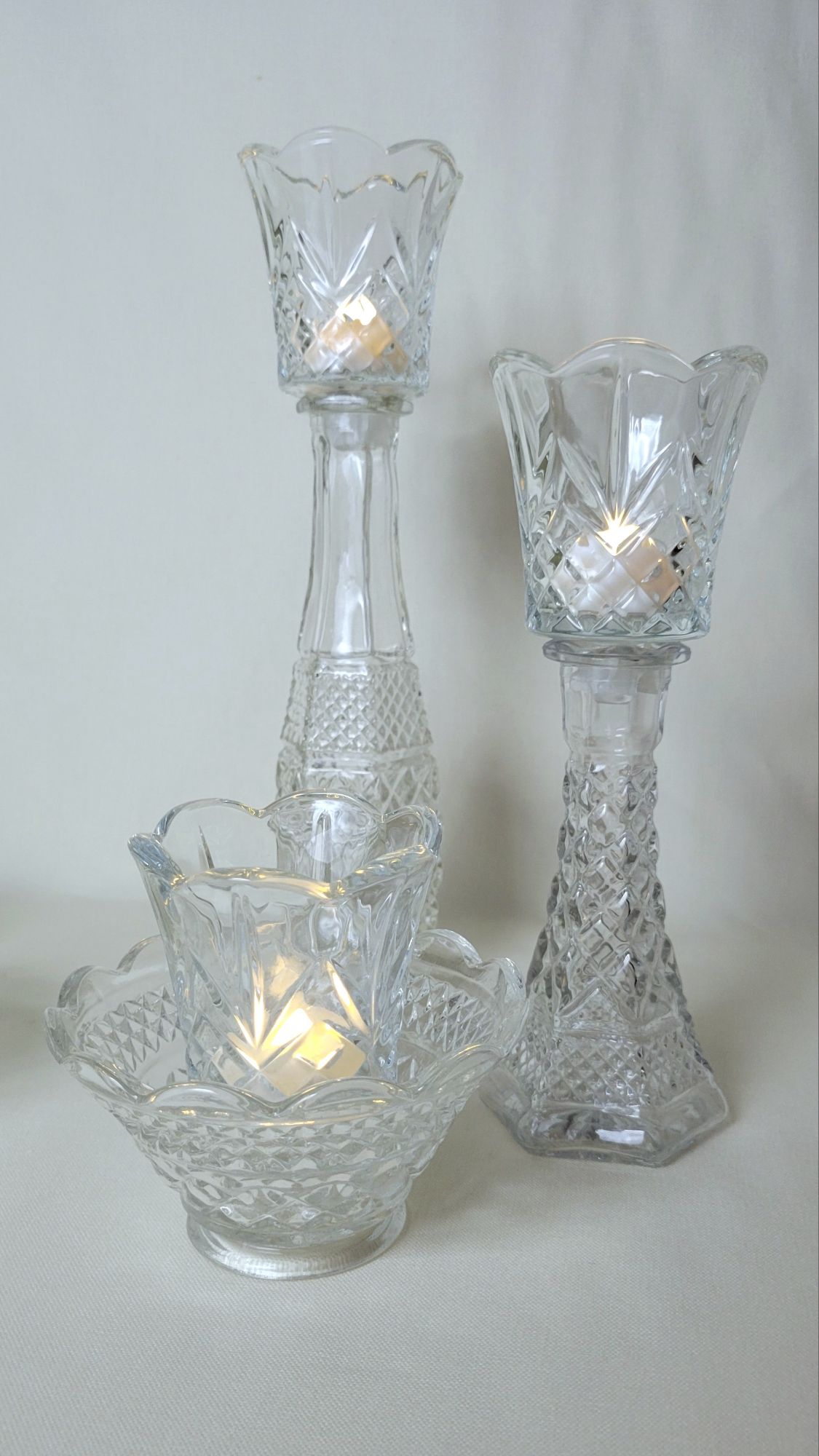 a grouping of Wexford pressed glass by Anchor Hocking, including a candle bowl, a six inch tall bud vase/candlestick, and a nine inch tall bud vase. all of them hold a Cambridge peg votive by HOMCO with an LED tealight.